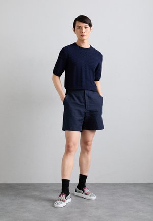 Pull en tricot à manches courtes à motif marine, shorts marins et chaussures à enfiler multicolores en zigzag portées avec des chaussettes noires.
