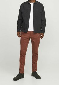 Svart skjorta med knappar och två bröstfickor, bärs över en vit t-shirt och matchas med rostfärgade slim-fit byxor och svarta skor.