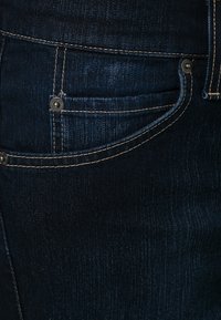 Mörkblå denimjeans med en framficka, synlig sömnad i ljusbrunt och metalldetaljer i knappform. Tyget har en slät textur.