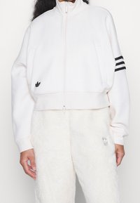 Valkoinen lyhyt takki, jossa on korkea kaulus, vetoketjusuljin, mustat kolmonauha-aksonnit hihoissa ja pieni musta logo vasemmassa rinnassa. Pehmeä materiaali.