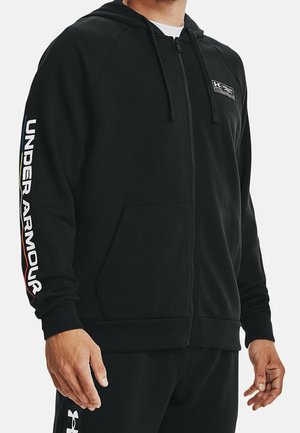 Mand iført sort Under Armour hættejakke med lynlås, logo på brystet og mærketekst med farvede striber på ærmet, kombineret med sorte bukser.