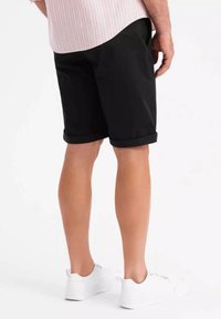 Shorts noirs avec des ourlets retroussés, en matériau lisse. Portés avec des baskets blanches. Un haut rayé rose clair est partiellement visible au-dessus.