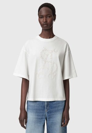 Kvinde iført en løs hvid t-shirt med korte ærmer og et tekstureret blomsterprint foran samt blå jeans, mod en ensfarvet baggrund.