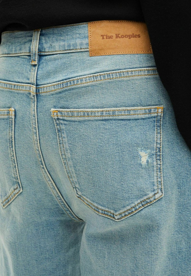 Light blue denim jeans with a sleek design, featuring a leather patch labeled "The Kooples" and subtle distressed detailing on the back.