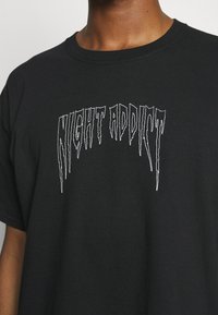 Černé bavlněné tričko s stylizovaným bílým grafickým textem "NIGHT ADDICT" v písmu podobném rampouchům napříč hrudníkem. Klasický design s kulatým výstřihem.