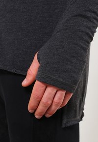 Hand, teilweise von Daumenloch in dunkelgrauem Langarmshirt bedeckt, ruht an schwarzer Hose.
