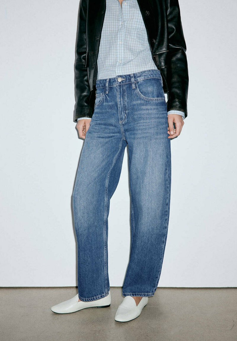 Personne portant un jean bleu droit, des chaussures blanches sans lacets, une chemise claire à carreaux et une veste en cuir noire, debout sur un sol gris.