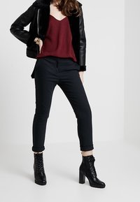Mujer vestida con chaqueta de cuero negra con cuello de piel, blusa burdeos de escote en V, pantalones ajustados negros y botines de tacón alto negros con cordones.