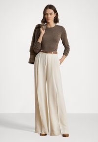Bruine blouse met lange mouwen, crème wijde broek, bruine riem met metalen gesp, en bruine hakken. Geruite blazer over de arm.