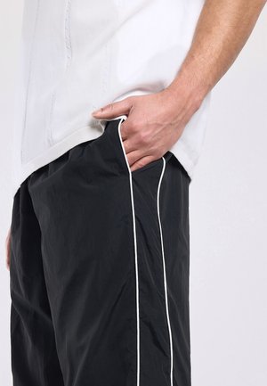 Persoon die zwarte broek draagt met witte strepen aan de zijkant en een wit overhemd, hand nonchalant in het voorvak van de broek gestoken.