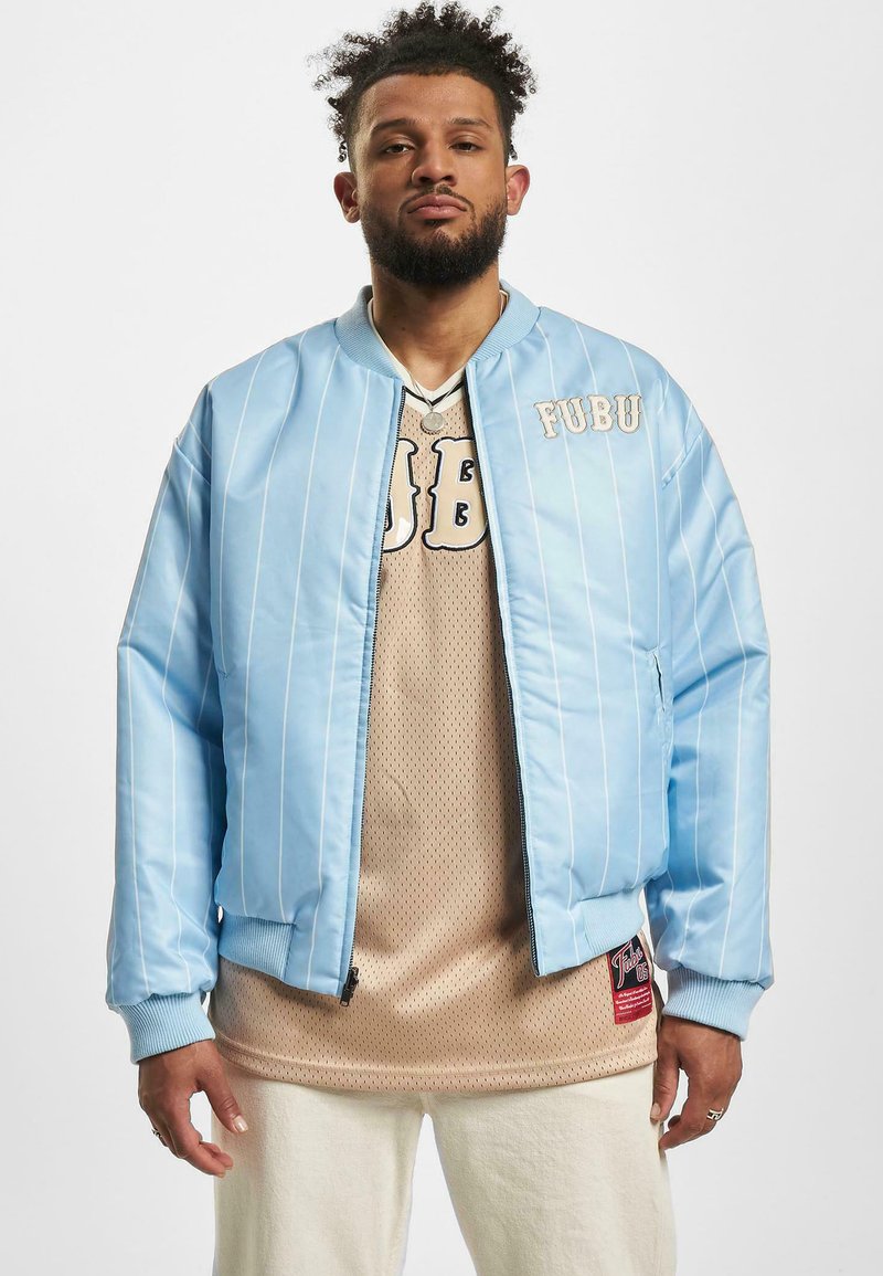 Veste bomber bleu clair avec des rayures blanches, un col et des poignets côtelés. Logo FUBU sur la poitrine. Chemise en mesh beige en dessous.
