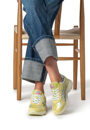 Zapatillas amarillas y grises con paneles texturizados, detalles de malla y cordones blancos. Usadas con jeans vaqueros remangados sobre una silla de madera.