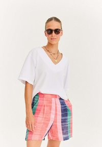 Žena nosící tmavé sluneční brýle, bílou blůzu s véčkovým výstřihem, barevné pruhované šortky a vrstvené korálkové náhrdelníky, stojící proti bílému pozadí.