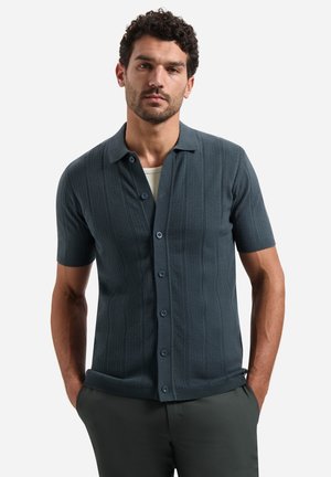 Mann mit kurzen lockigen Haaren und Bart, der ein dunkelblaues, strukturiertes Kurzarmhemd mit Knöpfen und dunkle Hosen trägt, die Hände in den Taschen.