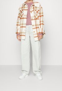 Karierte Flanellhemd in Creme, Braun und Orange mit zwei Brusttaschen. Darunter ein leichter, rosa Pullover. Hellblaue Jeans mit weitem Bein und weiße Sneakers.