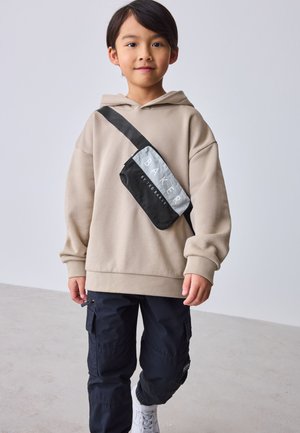 Młody chłopiec ubrany w beżową bluzę z kapturem, czarne spodnie typu cargo, białe tenisówki i czarną torbę na ramię z napisem „BAKER BY TED BAKER”.