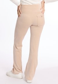 Pantalons de survêtement beiges évasés en tissu doux, dotés d'une poche arrière avec une étiquette et d'une taille haute, portés avec des baskets blanches.