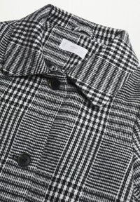 Černo-bílý kostkovaný kabát s límcem, s mřížkovaným vzorem a dvěma černými knoflíky na přední straně. Texturovaná látka.