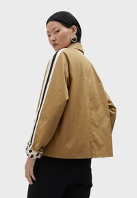 Giacca khaki con strisce laterali nere e bianche, con polsini a pois. Tessuto leggero e casual, design leggermente oversize.