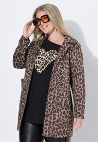 Leopardenmuster-Mantel aus braunem und schwarzem Stoff, mit einer Vordertasche, entspannter Passform und kombiniert mit einem schwarzen Shirt, das ein Leopardenherz-Design zeigt.