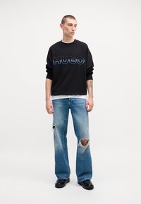 Felpa nera con logo "HUGO" in blu, abbinata a jeans chiari strappati e scarpe nere lucide. Design semplice e casual.