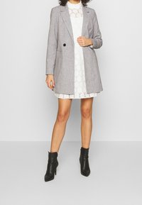 Szary żakiet o dopasowanym kroju, z pojedynczym czarnym guzikiem, w połączeniu z białą koronkową sukienką. Czarny skórzany botek się dopełnia stylizację.