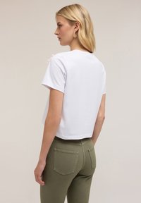T-shirt cropped bianco a maniche corte con un design pulito, abbinato a pantaloni aderenti verde oliva con tasche posteriori e una leggera texture.