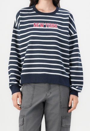 Persoon draagt een marineblauw-wit horizontaal gestreepte trui met de tekst "NEW YORK" in rood en wit, gecombineerd met grijze cargobroek.