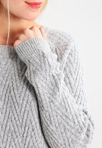 Femme portant un pull en maille texturée gris clair, tenant le col près du menton, souriant doucement devant un fond uni clair.