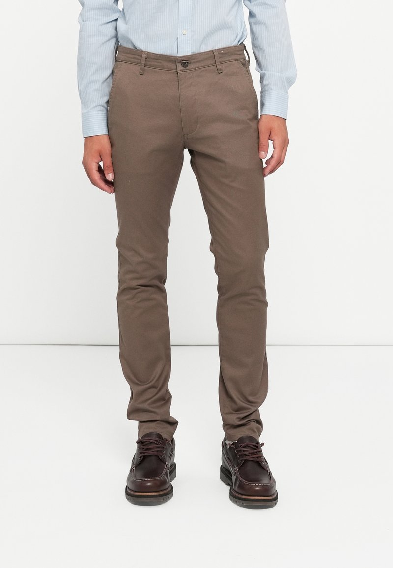 Homme portant un pantalon marron slim, des chaussures à lacets marron foncé et une chemise à rayures bleu clair, debout devant un fond blanc.