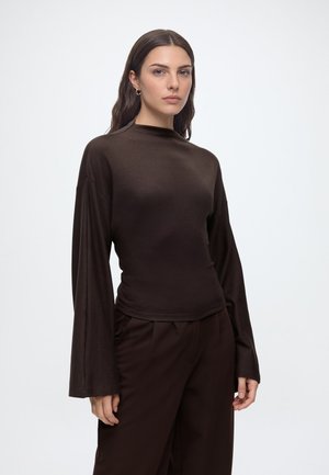 Femme aux longs cheveux foncés portant un haut marron foncé à manches larges et un pantalon assorti, debout devant un fond blanc uni.