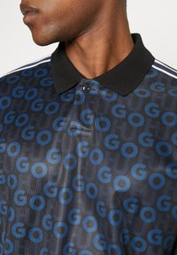 Personne portant un polo noir avec un col côtelé et un motif bleu "GO" sur le tissu.