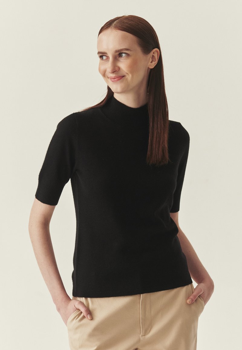 Pull noir à manches courtes avec un col montant, en matière douce. Le modèle porte un pantalon beige, les mains dans les poches, fond neutre.