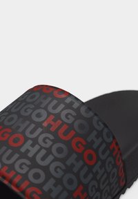 Sandale à glissière noire avec une large bride présentant un motif répétitif du logo « HUGO » en lettres rouges et gris foncé.