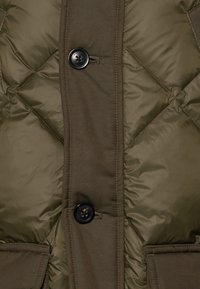 Chaleco acolchado color verde oliva con textura suave, que presenta un cierre de botones, un patrón de costura diagonal y bolsillos laterales.