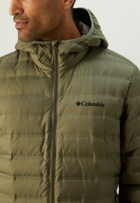 Olivenfarget isolert pufferjakke med glidelås og hette. Har horisontale quiltede mønstre og liten svart logo på brystet.