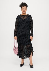 Ensemble composé de deux pièces en dentelle florale noire, comprenant un haut à manches longues et une jupe à volants. Le mannequin tient un sac à main en sequins rose. Des chaussures noires complètent la tenue.