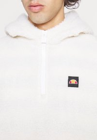 Vit fleecetröja med dragkedja och huva. Har en svart fyrkantig lapp med en färgglad logotyp. Mjuk textur med en mysig look.