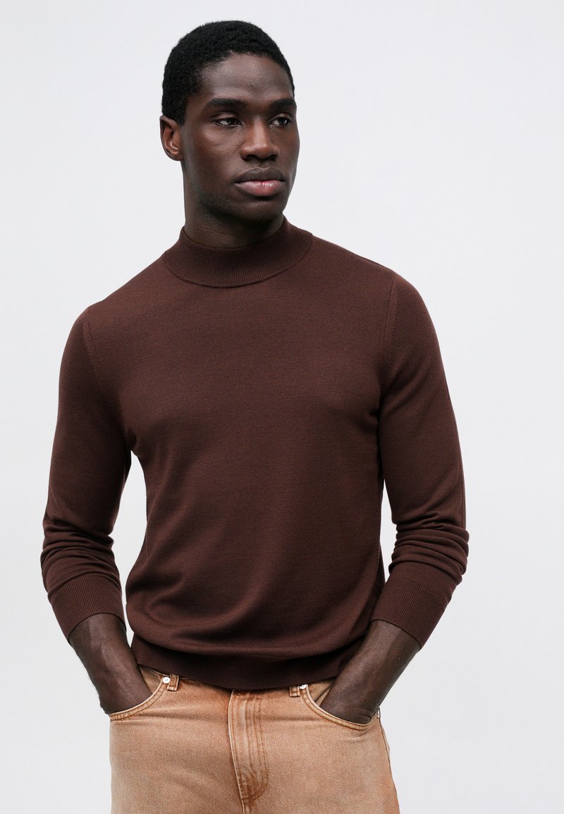 Brun ribbet sweater med høj hals, lange ærmer og en tætsiddende pasform, parret med lysebeige jeans.