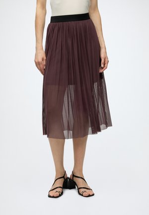 Femme portant une jupe midi plissée transparente de couleur violet foncé avec une ceinture noire, associée à des sandales noires à talons épais et fines brides.