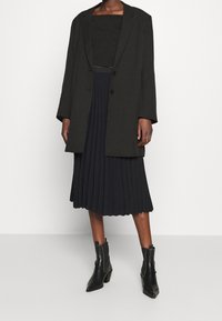 Blazer nero oversize, top nero corto e gonna midi plissettata nera abbinati a stivaletti con tacco neri. Texture lisce e design strutturato.