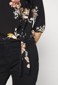 Blusa nera a fiori con maniche a tre quarti, con motivi floreali rosa e gialli, legata in vita. Indossata con pantaloni neri aderenti.