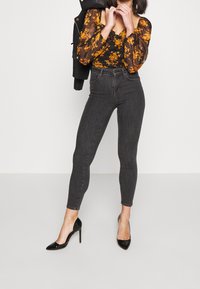 Jeans de mezclilla negros de tiro alto, con corte ajustado, que cuentan con bolsillos y una textura suave, combinados con una blusa de estampado floral y tacones negros.