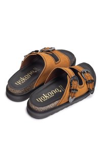 Sandales en suède marron avec deux boucles en métal noir, semelle texturée noire et détail en liège. Design facile à enfiler avec orteils et talons ouverts.
