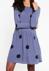 Pitkähihaista, polvenpituista hametta vaaleanliilasta kankaasta, jossa on mustia pilkkuja. Mekossa on istuva yläosa ja liehuvahelma.