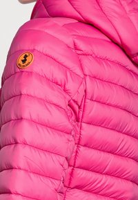 Giacca imbottita rosa con baffi orizzontali, dotata di un patch con logo e cappuccio. Il materiale appare liscio e lucido.