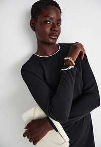 Μαύρο φόρεμα με μακριά μανίκια και λευκή λαιμόκοψη, συνδυασμένο με λευκή τσάντα. Σημαντικές βραχιόλια σε χρυσό και ασημένιο χρώμα στον καρπό.