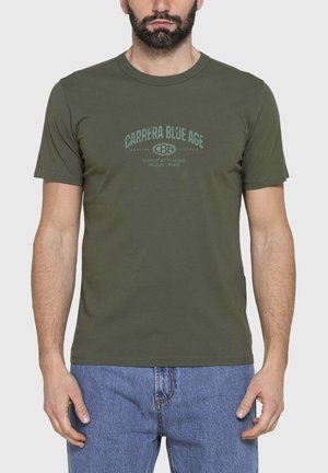Uomo che indossa una t-shirt verde oliva a maniche corte con scritta azzurra "CARRERA BLUE AGE" e jeans azzurri, in piedi su uno sfondo uniforme.