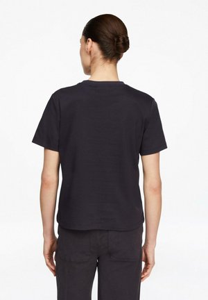 Kobieta z brązowymi włosami upiętymi w kok, ubrana w prostą czarną koszulkę z krótkim rękawem i czarne spodnie, stojąca tyłem na białym tle.