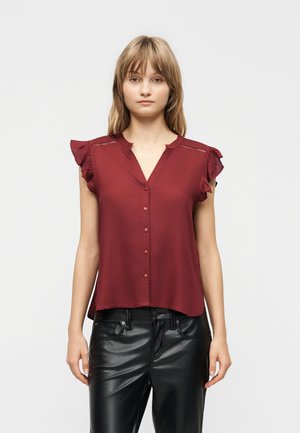 Woman wearing a sleeveless burgundy button-up blouse with ruffled shoulders and black leather pants, standing against a plain light background.